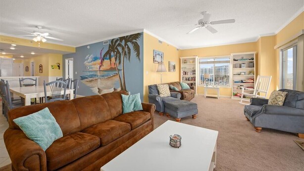 A living room with a brown leather sofa, a blue armchair, and a white coffee table. There is a painting of a beach scene on the wall, a ceiling fan, and a bookshelf filled with books. - 3 br oceanview 45th St with pool (Ocean City)