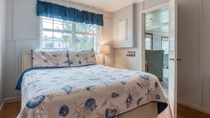 A bedroom with a bed, a window with a view of houses, a lamp, and a door leading to another room. - Perfect Beach Cottage, Friendly, Walk to Beach and Seaside Promenade (Seaside)