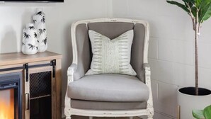 A living room with a grey armchair, a flat-screen TV, a fireplace, a framed picture, and a potted plant. - Cozy Modern Studio Just 2 Blocks To Forsyth Park! (Savannah)