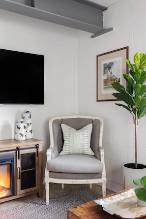 A living room with a grey armchair, a flat-screen TV, a fireplace, and a framed picture on the wall. - Cozy Modern Studio Just 2 Blocks To Forsyth Park! (Savannah)