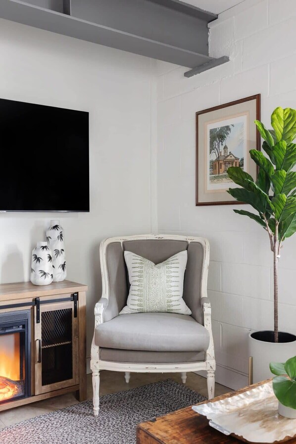 Una sala de estar con un sillón gris, un televisor de pantalla plana, una chimenea, un cuadro enmarcado y una planta en maceta.