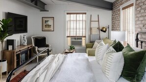 A modern hotel room with a bed, a desk, a chair, a plant, and a window with blinds. - Cozy Modern Studio Just 2 Blocks To Forsyth Park! (Savannah)