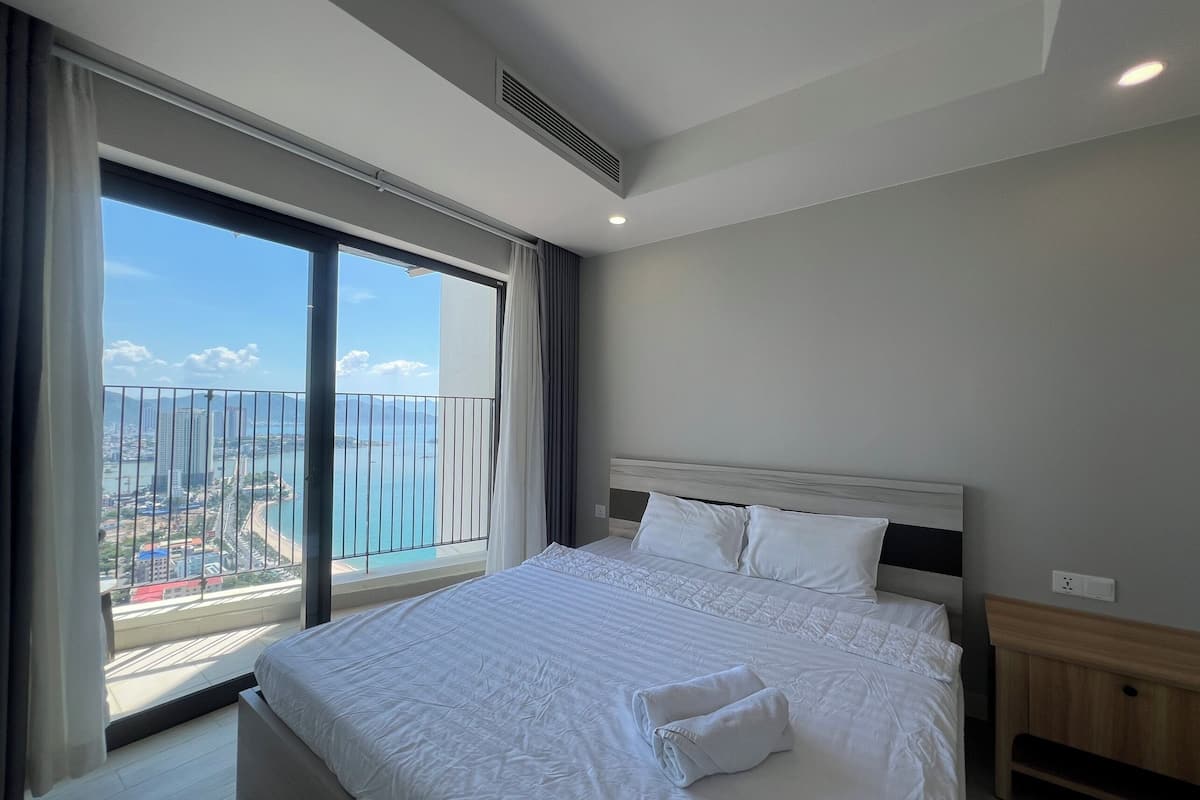 Een hotelkamer met een groot bed, een houten nachtkastje en een uitzicht op een stadsgezicht vanaf een balkon.