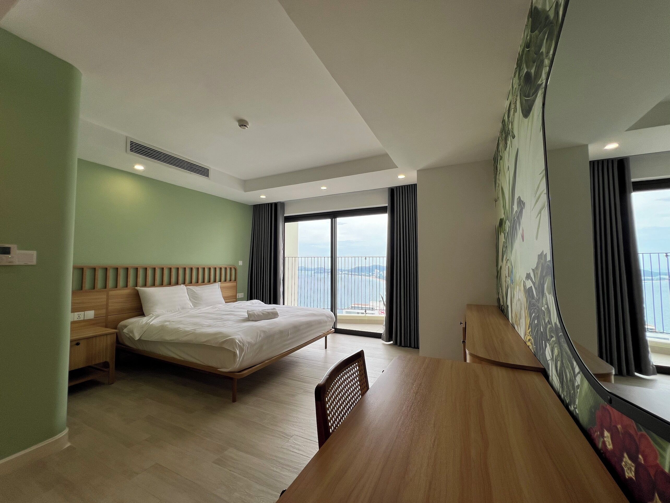 A hotel room with a bed, a desk, a chair, a large window with a view, and a painting on the wall.