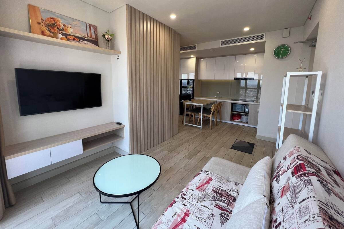 Een moderne woonkamer met een flatscreen-tv, een klein rond tafeltje, een bank met opvallende kussens en een keuken op de achtergrond.