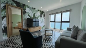 A living room with a large TV on a wooden stand, a sofa, a chair, and a view of the sea through a large window.