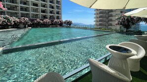 A swimming pool with clear water, surrounded by wicker chairs and umbrellas, with a view of a high-rise building and a landscaped area with pink flowers.