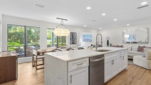 A modern kitchen with a central island, white cabinetry, and a large window overlooking a garden. - Charming Coastal 2BR Villa, GOLF CART, Cozy Sunroom, Near Beach, Pool, Wharf. (Miramar Beach)