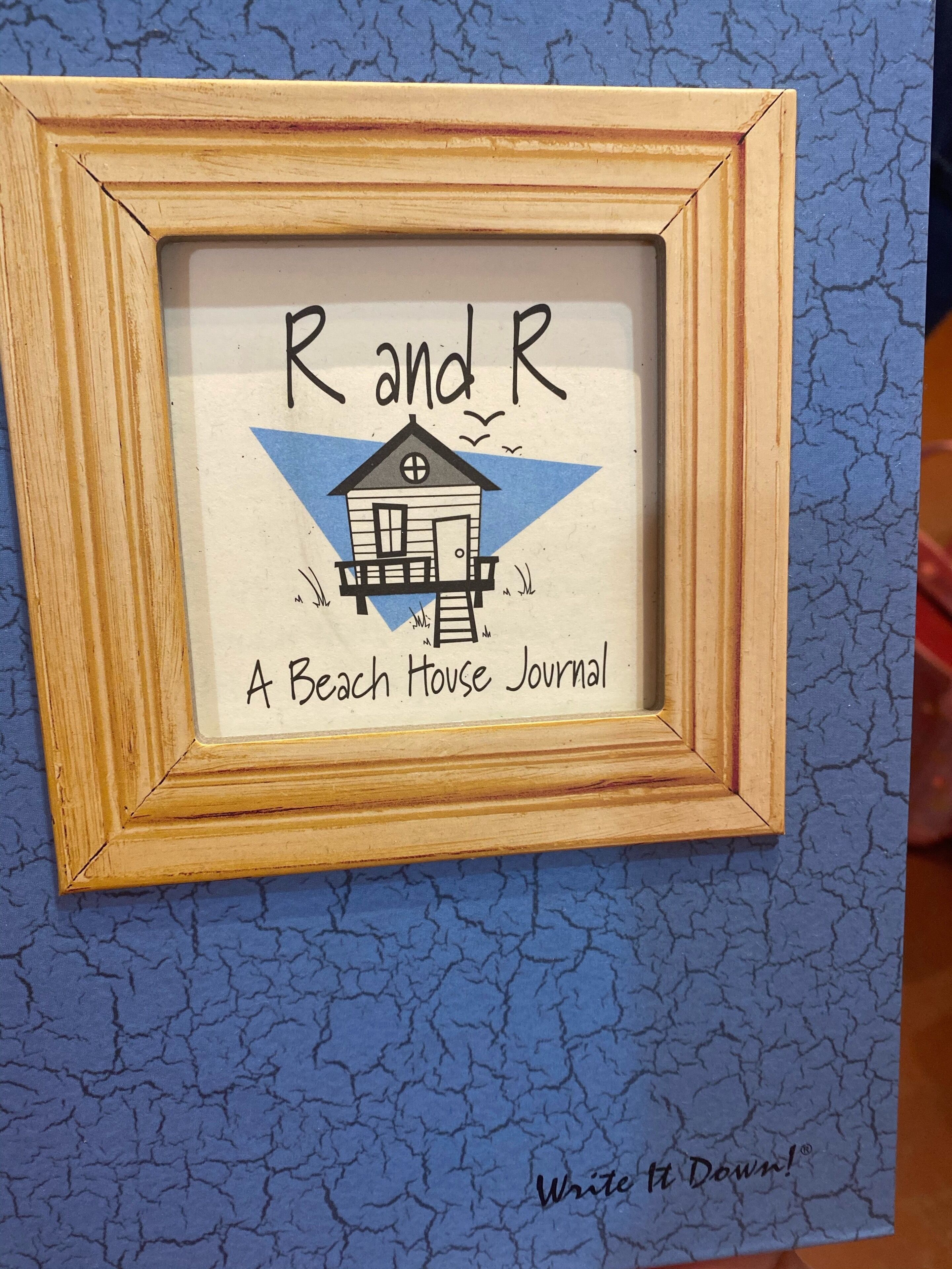 Ein gerahmtes Kunstwerk mit einer Hausillustration und dem Text 'Rand R A Beach House Journal' und 'Write It Down!' vor einem blauen Hintergrund mit Struktur.