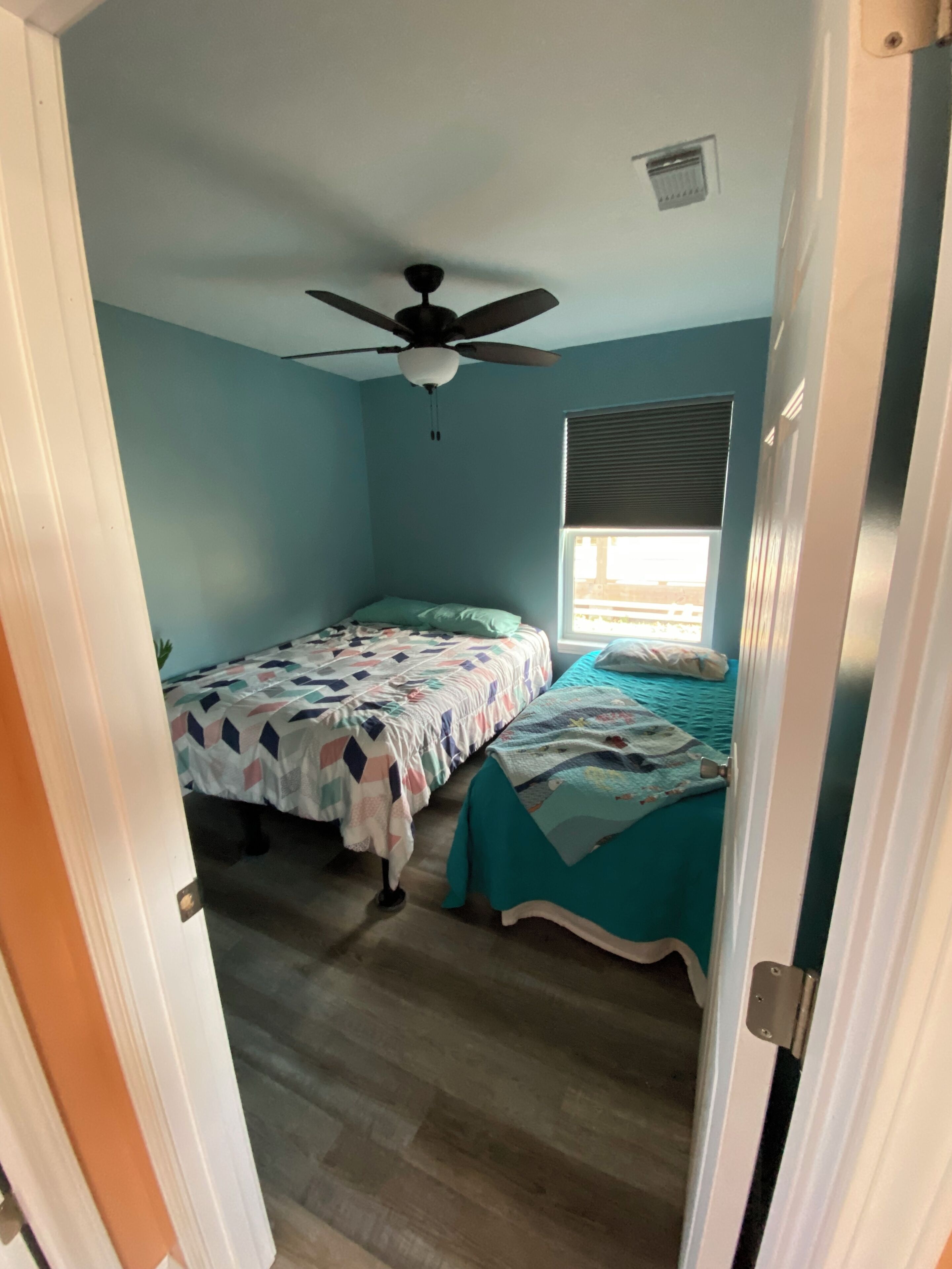 Une chambre avec deux lits, un ventilateur de plafond, une fenêtre avec des stores et une porte.