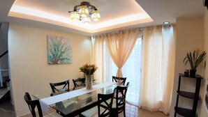 A dining area with a glass table, black chairs, a vase with plants, a painting on the wall, and a ceiling light fixture.