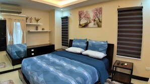 A bedroom with a bed, a nightstand, a window with blinds, a wall-mounted air conditioner, a shelf with decorative items, and a painting on the wall.