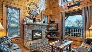 Ein gemütliches Blockhaus-Interieur mit Kamin aus Naturstein, Holzmöbeln und einem herrlichen Ausblick durch ein großes Fenster.