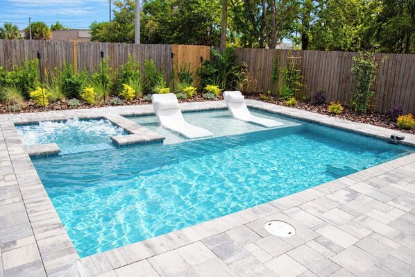 A rectangular swimming pool with three white lounge chairs and a wooden fence in the background.