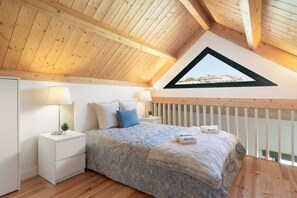 Un dormitorio con techo inclinado, una cama con colcha de diseño azul y blanco, dos mesitas de noche con lámparas y vistas a través de un ventanal triangular.