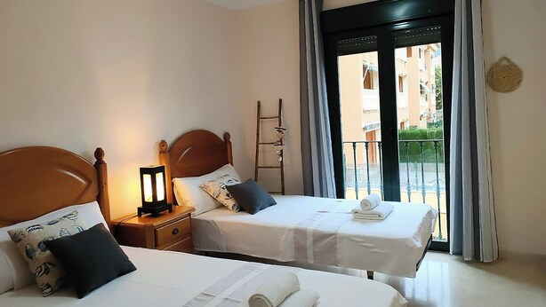 A hotel room with two beds, each with a pillow, a nightstand, a lamp, and a view of the outside balcony. - Holiday Apartment - Javea Puerto - 2bedr 2 Bath (Javea)