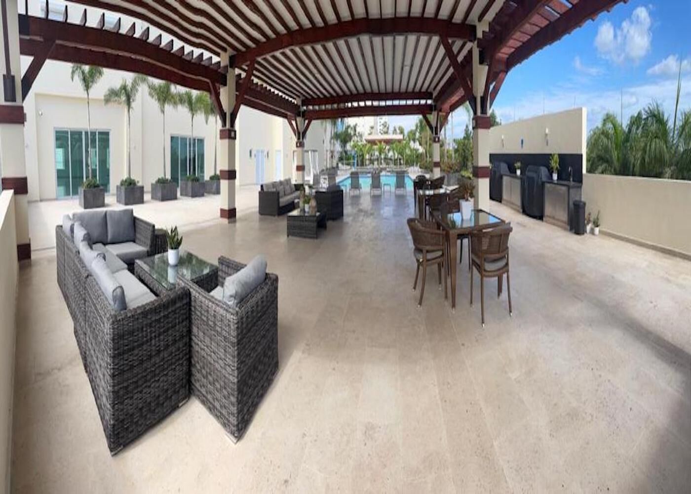 A covered outdoor area with wicker furniture, a pool, and a view of a building with palm trees.