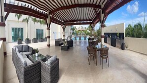 Un espacio al aire libre techado con muebles de mimbre, piscina y vistas a un edificio con palmeras.