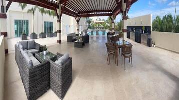 A covered outdoor area with wicker furniture, a pool, and a view of a building with palm trees.
