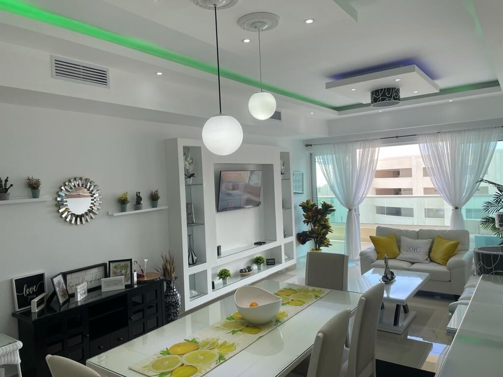 Una sala de estar moderna con un sofá blanco, una mesa de comedor con un cuenco y un televisor en la pared.