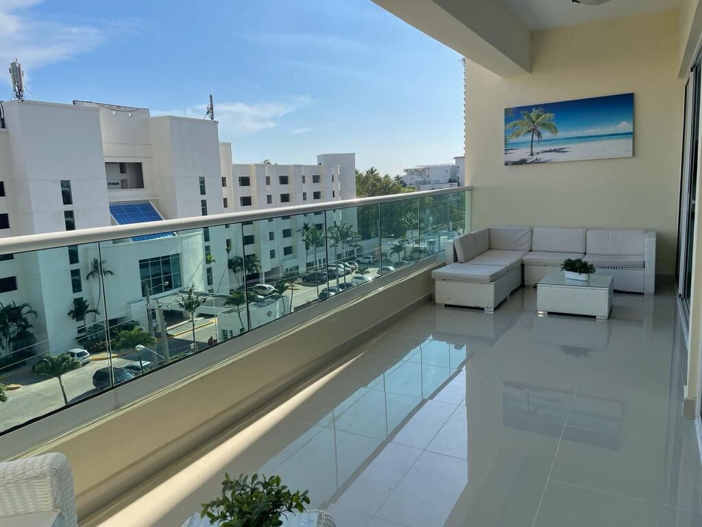 Un balcón moderno con barandilla de vidrio, muebles blancos y vistas a edificios y palmeras.
