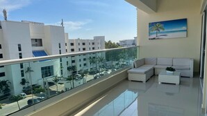 Un balcón moderno con barandilla de vidrio, muebles blancos y vistas a edificios y palmeras.