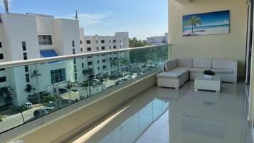Un balcón moderno con barandilla de vidrio, muebles blancos y vistas a edificios y palmeras.
