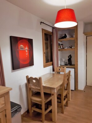 Un comedor con una mesa y sillas de madera, una lámpara colgante roja, un cuadro en la pared y una zona de cocina con un refrigerador y estanterías.