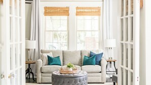 Ein Wohnzimmer mit einem beigen Sofa, türkisfarbenen Kissen, einem grauen Hocker und einem Korbwannenmöbel.