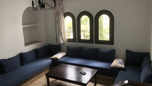 Een kamer met een donker houten salontafel, een blauwe bank met gestreepte kussens, en drie gewelfde ramen die natuurlijk licht binnenlaten.