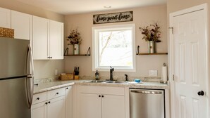 A kitchen with white cabinets, a stainless steel refrigerator, a dishwasher, and a window with a flower arrangement.