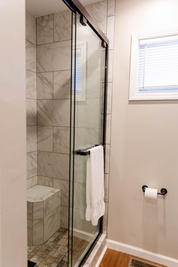Ducha con paredes de vidrio, azulejos de mármol, una toalla blanca y un dispensador de papel higiénico en la pared.