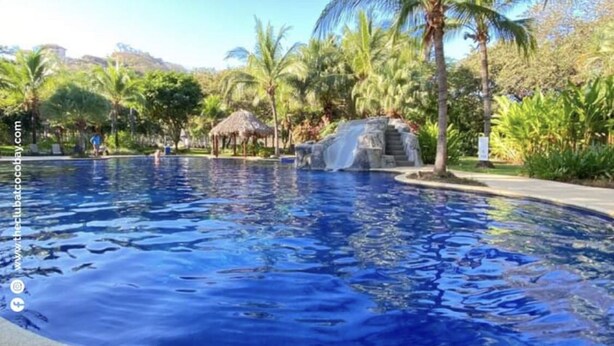 Ein Pool mit Wasserfall, umgeben von Palmen und einer Struktur mit einem Reetdach.