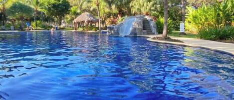 A swimming pool with a waterfall feature, surrounded by palm trees and a thatched-roof structure.