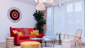 Una sala de estar moderna con un sofá rojo, una mesa de centro redonda de madera, un taburete amarillo, una silla rosa y una planta grande en maceta.
