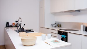 Una cocina moderna con armarios blancos, una campana extractora de acero inoxidable, horno integrado y encimera con tabla de cortar, cuencos y cafetera.