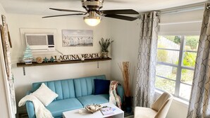 Una sala de estar con un sofá azul, una mesa de centro blanca, un sillón beige, un ventilador de techo, un ventanal con cortinas estampadas y una estantería con objetos decorativos.