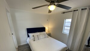 Un dormitorio con cama, ventilador de techo, mesita de noche con lámpara y ventana con cortinas.