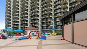 A multi-story residential building with balconies, a children's play area with slides and a water feature, and outdoor seating under umbrellas.