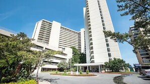 A modern high-rise building with multiple floors, surrounded by greenery and a parking area.