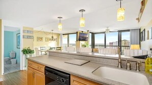 A modern kitchen with a large island, a dishwasher, and a sink. There are pendant lights above the island and a large window providing a view of the city.