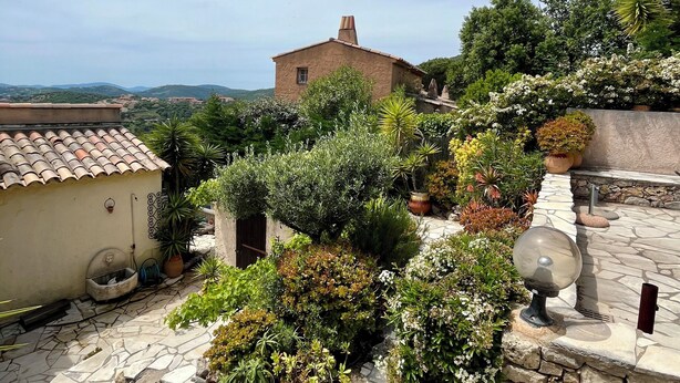 Une cour dallée de pierres, avec un bâtiment rustique, une végétation luxuriante et un ciel dégagé.