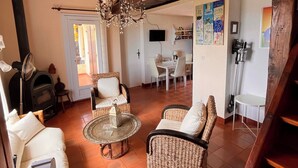 Un vaste espace de vie doté d’un haut plafond, de poutres apparentes, d’un lustre, de meubles en osier, d’une cheminée et d’un coin repas avec des chaises et une table.