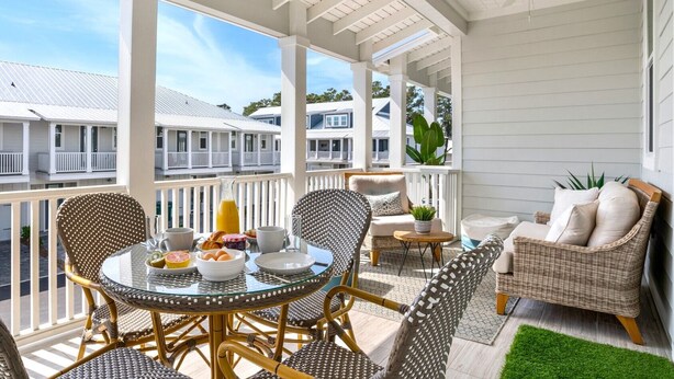 A covered outdoor seating area with wicker furniture, a glass table, and a view of neighboring buildings. - Golf Cart - 2 min to Beach - Pool - Walk to Shops & Dining! (Santa Rosa Beach)