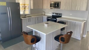 A modern kitchen with light wood cabinets, stainless steel appliances, and a central island with two bar stools. - Modern, chic waterfront 3-bedroom villa w/ pool (Saint Ann's Bay)