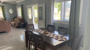 A dining area with a wooden table, chairs, and a quilt runner. There are two doors leading to an outdoor area with greenery visible through the windows. - Modern, chic waterfront 3-bedroom villa w/ pool (Saint Ann's Bay)