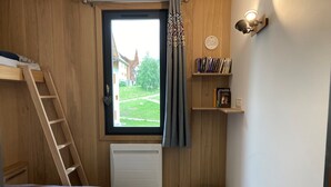 Ein Schlafzimmer mit Fenster, einer Leiter, einem Heizkörper, einem Nachttisch, einem grünen Ottomanen und einem Bücherregal.