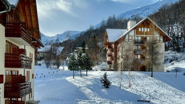 Ein verschneites Dorf mit Holzhäusern, eine Skipiste im Hintergrund und Berge in der Ferne.