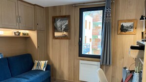 Ein gemütliches Zimmer mit einem blauen Sofa, einem weißen Sessel, Holzwänden, einem Fenster mit Blick auf Gebäude und einer Heizung.