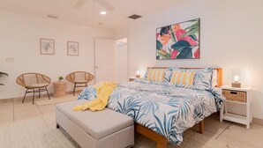 A bedroom with a bed, two chairs, a bedside table with a lamp, a ceiling fan, and a wall with a painting.